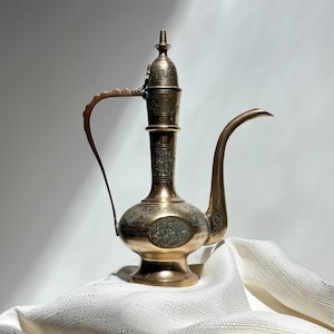 May include: A vintage brass water pitcher with intricate floral engravings. The pitcher features a long, curved spout, a handle, and a decorative lid. The brass has a warm, golden hue, and the design suggests an antique or Middle Eastern origin.