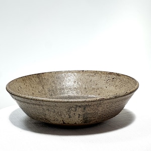 May include: A shallow, wide-rimmed ceramic bowl featuring a speckled, earthy beige and brown glaze. The exterior surface displays a textured, mottled finish with darker brown flecks distributed across the light tan base, creating a rustic appearance.