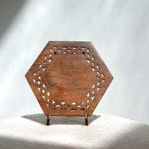 May include: A hexagonal wooden tray with intricate carved openwork around the edges. The tray is a warm brown color and is supported by small, gold-colored metal legs. The design is symmetrical and geometric, suitable for decorative use.