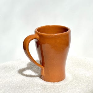 May include: A handmade, brown ceramic mug with a curved handle. The mug has a smooth, glossy finish and a slightly tapered shape. The mug is approximately 4 inches tall and is suitable for hot or cold beverages.