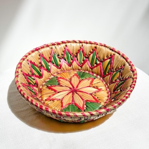 May include: A round woven basket with a floral design in the center. The basket is made of natural-colored straw with red, green, and pink accents. The rim is trimmed with red. The basket is likely used for decorative or storage purposes.
