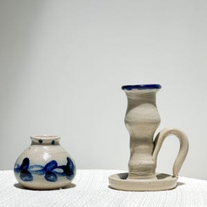 May include: A small, round, beige ceramic vase with a blue leaf pattern and a matching beige ceramic candlestick holder featuring a blue rim and a curved handle. Both items have a speckled texture and a glossy finish, presented together.