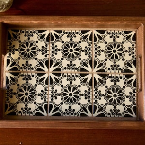May include: A wooden serving tray with a decorative tile insert featuring a black and white floral pattern. The tray is rectangular and has a dark wood frame. Decorative glassware and ornaments are visible.