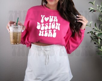 Puede incluir: Una sudadera rosa de cuello redondo, corta, con el texto "YOUR DESIGN HERE" en blanco y un corazón en el medio de la palabra "YOUR". La modelo lleva una falda blanca.