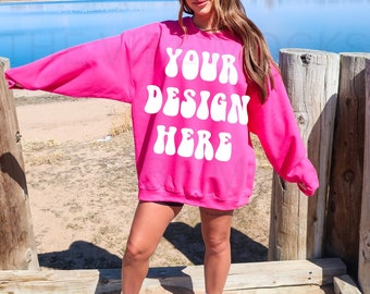 Puede incluir: Una sudadera rosa con el texto "YOUR DESIGN HERE" en blanco, con letras de estilo retro. La sudadera es de gran tamaño y tiene mangas largas.