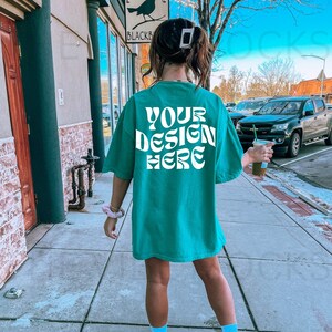 May include: Teal oversized t-shirt with the text "YOUR DESIGN HERE" in white, displayed on the back. The shirt is worn outdoors on a sunny day.