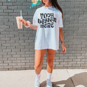 May include: A white oversized t-shirt with the text "YOUR DESIGN HERE" printed in black. The shirt is being worn by a person standing in front of a brick wall.