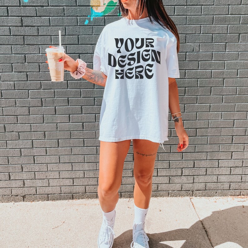 May include: A white oversized t-shirt with the text "YOUR DESIGN HERE" printed in black. The shirt is being worn by a person standing in front of a brick wall.