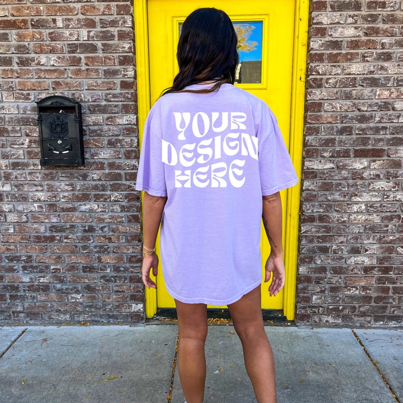 Op de afbeelding: Een lavendelkleurig oversized T-shirt met de tekst "YOUR DESIGN HERE" in wit op de achterkant. Het shirt wordt gedragen door een persoon die voor een gele deur staat.