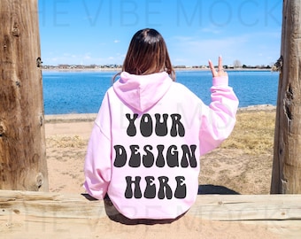 Puede incluir: Una sudadera con capucha rosa con el texto "YOUR DESIGN HERE" en letras negras de estilo retro. La sudadera con capucha la lleva puesta una persona de pie al aire libre cerca de una masa de agua.