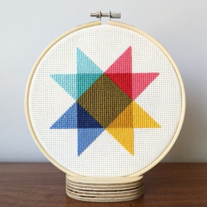 May include: A colorful geometric star pattern cross-stitched on white fabric, framed in a wooden hoop and displayed on a wooden stand.