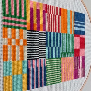 May include: A colorful cross-stitch embroidery in a wooden hoop. The design features a patchwork of geometric patterns in various colors, including orange, pink, green, blue, and black. The stitches are neatly aligned, creating a textured surface.