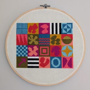 May include: A colorful geometric pattern cross-stitch design in a wooden embroidery hoop. The design features a variety of shapes and colors, including pink, blue, green, yellow, and black.