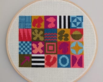 May include: A colorful geometric pattern cross-stitch design in a wooden embroidery hoop. The design features a variety of shapes and colors, including pink, blue, green, yellow, and black.
