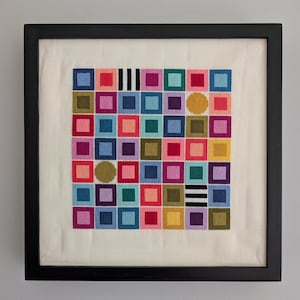 May include: A framed cross-stitch embroidery featuring a grid of colorful squares and circles on off-white fabric. The design includes various shades of blue, pink, red, yellow, purple, green, and black-and-white striped patterns within a black frame.