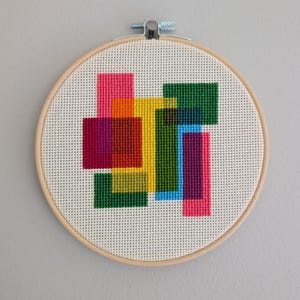 May include: A cross-stitch design with overlapping geometric shapes in pink, orange, yellow, green, blue, and purple. The artwork is displayed in a round wooden embroidery hoop, creating a modern, abstract wall hanging.
