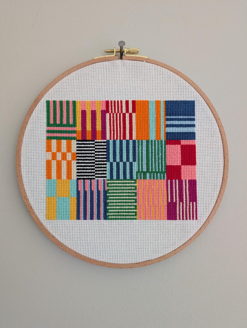 May include: A colorful cross-stitch design featuring a grid of geometric patterns in various colors, including pink, orange, blue, and green. The artwork is displayed in a round wooden embroidery hoop, creating a framed piece of textile art.