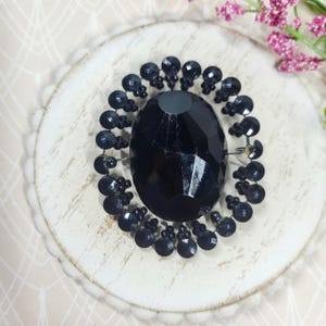 May include: A circular brooch featuring a large, central, faceted black oval stone surrounded by a ring of smaller, round, black faceted stones. The design is symmetrical, with the smaller stones arranged in a repeating pattern around the centre.