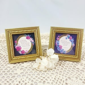 May include: Two square gold-toned frames with ornate detailing, each containing a dark blue background with a circular floral border. The left frame displays the text Love the Lord your God with all your heart and all your soul and all your strength. Deuteronomy 6:5. The right frame displays the text But as for me and my household, we will serve the Lord. Joshua 24:15.