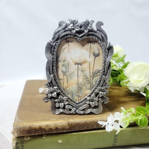 May include: A heart-shaped picture frame with a metallic silver finish and ornate, raised floral detailing along the edges. The frame contains an insert featuring a botanical illustration of white poppy flowers and buds against a beige background.