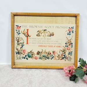 May include: A rectangular framed print titled The Brownie Scout Promise, featuring illustrations of small elf-like figures, floral vines, and text. The print is set within a light brown bamboo-style frame with a textured cream-colored background.