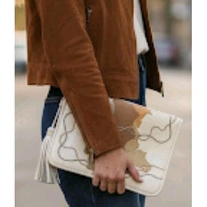 May include: A brown suede jacket worn with dark blue denim jeans and a white rectangular clutch bag. The clutch features a tan and white abstract pattern with thin dark lines and a long white tassel attached to the side zipper pull.