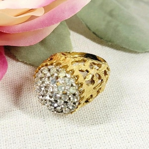 Puede incluir: Un anillo de tono dorado con una banda texturizada de aspecto orgánico y diseño calado. El centro del anillo es un grupo redondeado y elevado, densamente engastado con numerosas piedras pequeñas, claras y brillantes que captan la luz.