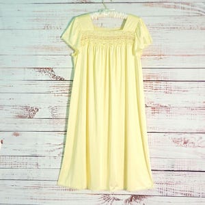 May include: A pale yellow nightgown featuring short flutter sleeves, a square neckline, and a smocked bodice with intricate white embroidery detailing. The garment has a loose, flowing skirt that falls to a knee-length hemline.