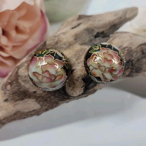 Puede incluir: Un par de pendientes redondos con un diseño floral. Los pendientes presentan un fondo negro con un diseño de flores rosas y blancas, delineado en oro. Los pendientes están montados sobre una base de metal dorado.