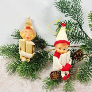 May include: Two vintage elf ornaments with painted facial features. One elf wears a gold hat and gold outfit, while the other wears a white hat with a red band, a red scarf, and white clothing. Both figures have arms crossed and are posed in a seated position.