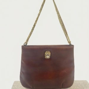 May include: A brown leather handbag with a smooth finish and a rectangular shape. The bag features a gold-toned metal chain strap and a central gold-toned metal emblem depicting a lion head on the front exterior of the leather body.
