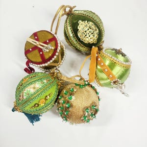 May include: Five decorative spherical ornaments featuring varied textures and colors. The collection includes green velvet, gold fabric, and satin finishes adorned with intricate gold trim, white pearl beads, sequins, and metallic embellishments with hanging loops.