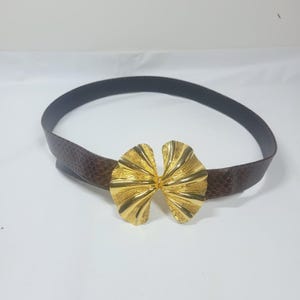 Puede incluir: Un cinturón marrón con un patrón de piel de serpiente texturizado y una hebilla dorada en forma de lazo. El cinturón mide aproximadamente 2,5 cm de ancho y se muestra sobre un fondo blanco. La hebilla tiene un diseño acanalado.