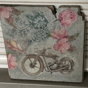 May include: A vintage-style wooden sign with a floral design and a motorcycle illustration. The sign features pink flowers, green leaves, and a gray motorcycle with a brown leather seat.