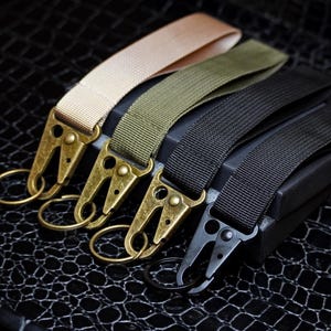 Op de afbeelding: Vier sleutelhangers met verschillende gekleurde bandjes: beige, olijfgroen en zwart. Elke sleutelhanger heeft een metalen sluiting en een kleine metalen ring. De sluitingen zijn goud en zwart. De sleutelhangers liggen op een zwart oppervlak met textuur.