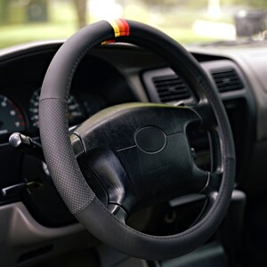 May include: A black leather steering wheel cover with red, yellow, and black accents. The cover has a perforated design and is installed on a black steering wheel.
