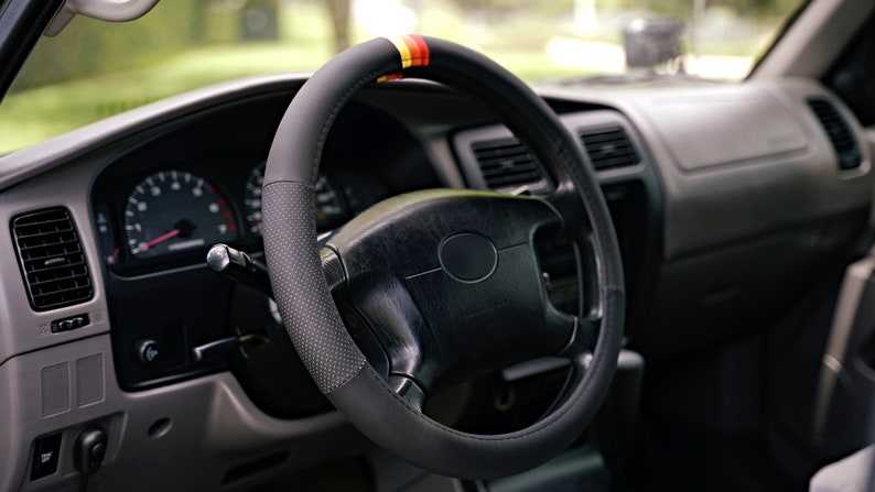 May include: A black leather steering wheel cover with red, yellow, and black accents. The cover has a perforated design and is installed on a black steering wheel.