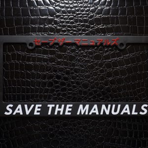 Könnte beinhalten: Schwarzer Nummernschildrahmen mit weißem Text "SAVE THE MANUALS" und rotem japanischem Text "セーブ・ザ・マニュアルズ".