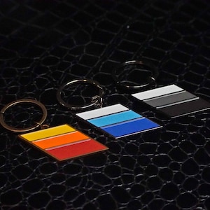 Puede incluir: Tres llaveros con rayas de diferentes colores. El primer llavero tiene rayas rojas, naranjas y amarillas. El segundo llavero tiene rayas blancas, azules y azul claro. El tercer llavero tiene rayas negras, grises y gris oscuro.