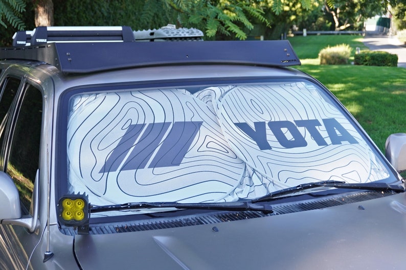 Pu&ograve; includere: Un parasole per parabrezza bianco con un motivo a mappa topografica nera e il testo YOTA stampato in grassetto nero. Il parasole &egrave; posizionato all'interno del parabrezza anteriore di un veicolo per bloccare la luce solare.