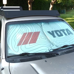 Pu&ograve; includere: Un parasole per parabrezza azzurro con un motivo a linee topografiche nere, una grafica a tre strisce rosse sulla sinistra e la parola YOTA stampata in grassetto nero in maiuscolo sulla destra, installato all'interno di un veicolo argento.
