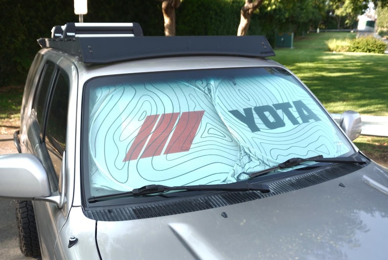 Pu&ograve; includere: Un parasole per parabrezza azzurro con un motivo a linee topografiche nere, una grafica a tre strisce rosse sulla sinistra e la parola YOTA stampata in grassetto nero in maiuscolo sulla destra, installato all'interno di un veicolo argento.