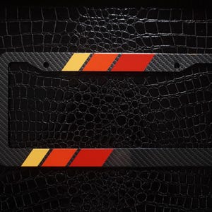 Puede incluir: Un marco de matrícula rectangular con una textura de fibra de carbono negra. El marco está acentuado con dos conjuntos de rayas diagonales en amarillo, naranja y rojo, situadas en la parte superior central y en la esquina inferior izquierda.