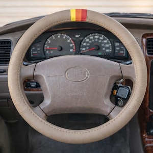 Puede incluir: Funda de volante beige con diseño perforado. La parte superior del volante tiene una franja amarilla, naranja y roja. El salpicadero presenta indicadores, un reloj digital y una pantalla de mapa. El interior es marrón y beige.