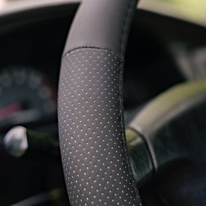 May include: Close-up of a black leather steering wheel cover with a perforated pattern. The cover is designed to improve grip and comfort.
