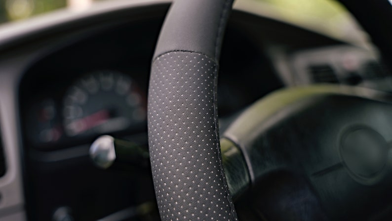 May include: Close-up of a black leather steering wheel cover with a perforated pattern. The cover is designed to improve grip and comfort.