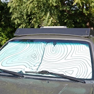 Pu&ograve; includere: Un parasole per parabrezza di veicolo con sfondo bianco e un motivo a linee di mappa topografica nere. Il parasole &egrave; posizionato all'interno del parabrezza anteriore di un veicolo grigio, dotato di un portapacchi nero sul tetto.