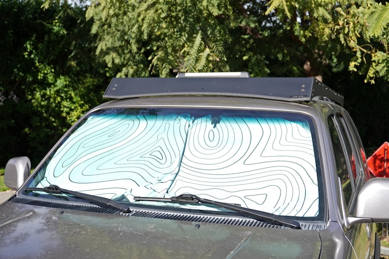 Pu&ograve; includere: Un parasole per parabrezza di veicolo con sfondo bianco e un motivo a linee di mappa topografica nere. Il parasole &egrave; posizionato all'interno del parabrezza anteriore di un veicolo grigio, dotato di un portapacchi nero sul tetto.