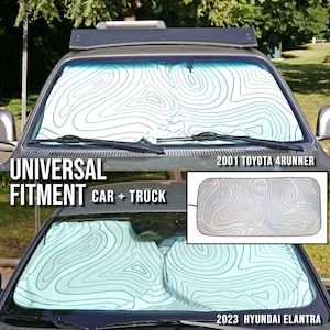 Pu&ograve; includere: Un parasole per parabrezza con sfondo bianco e linee di contorno topografiche nere. Il parasole &egrave; installato su una Toyota 4Runner del 2001 e una Hyundai Elantra del 2023, con il testo Universal Fitment Car + Truck.