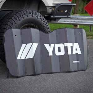 May include: A black and gray sunshade with the word "YOTA" in large white letters. The sunshade has a striped pattern and a white logo on the left side. The sunshade is leaning against a vehicle tire.